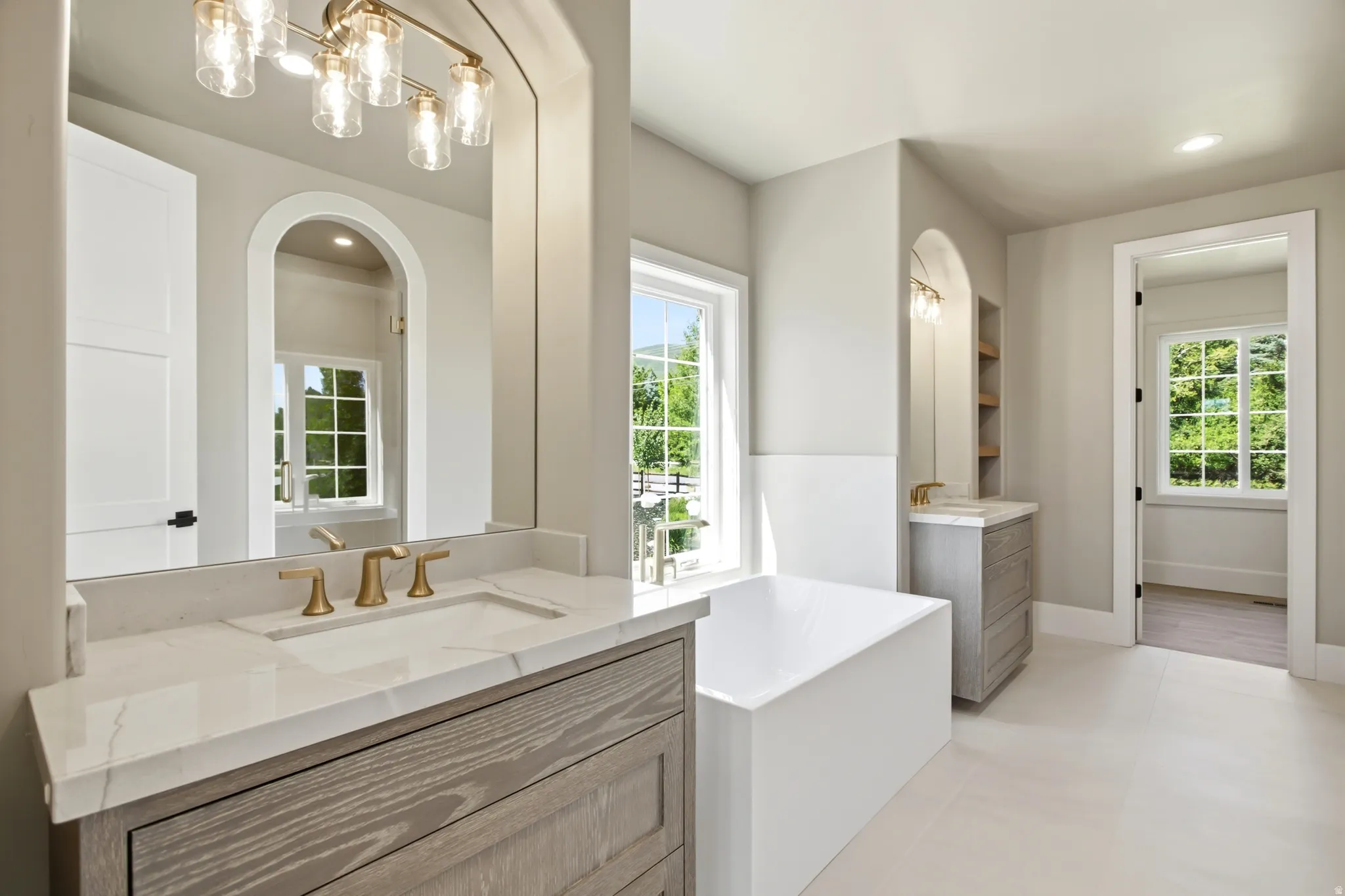 Contemporary primary bathroom featuring a freestanding tub, dual vanity with light wood-finish cabinetry, white countertops, and brass fixtures