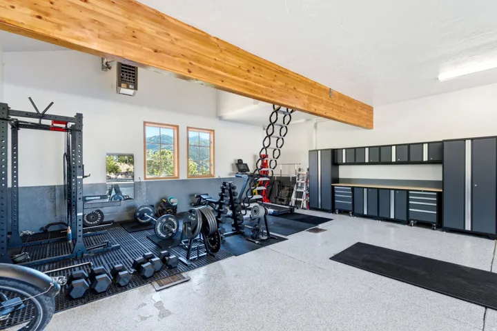 Spacious garage featuring a light wood ceiling beam, light gray speckled epoxy flooring, built-in gray cabinetry with a wood-finish countertop, and multiple windows