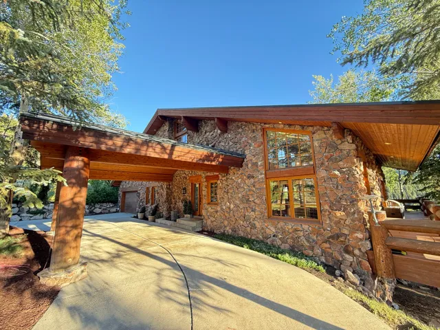 Stone exterior residence featuring a porte-cochère with log supports, extensive wood trim, large multi-pane windows, and a paved driveway