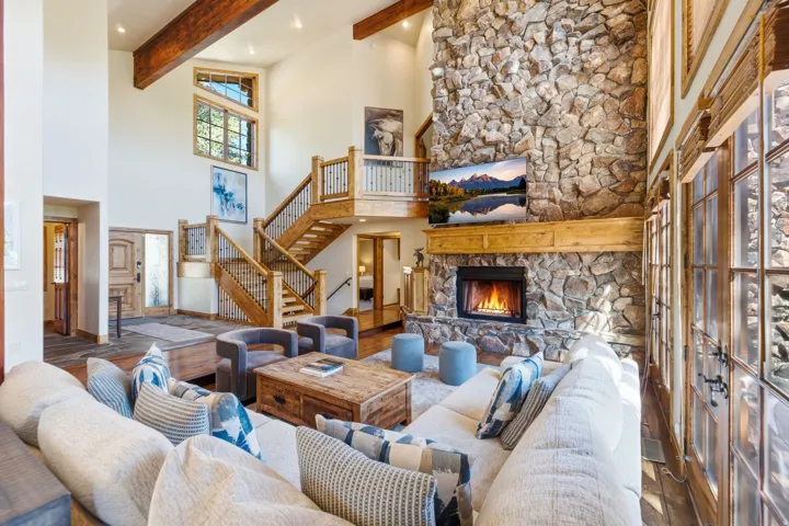 Spacious living area featuring a stone fireplace with a wood mantle, high ceilings with exposed wood beams, and large multi-pane windows
