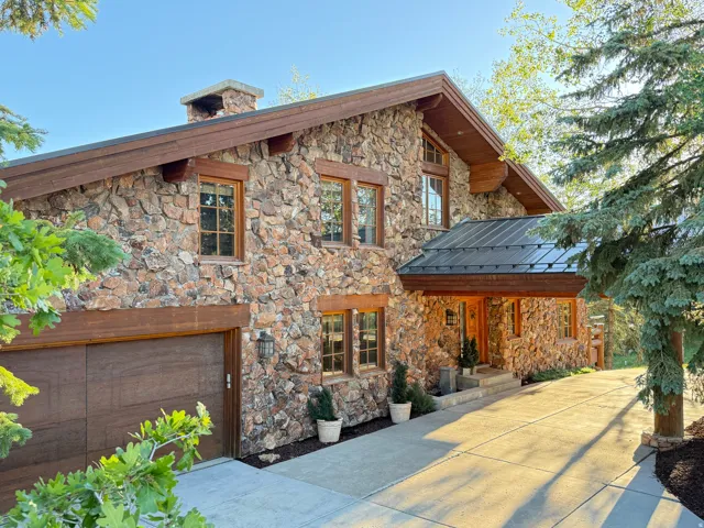 Stone-clad exterior featuring a wood-finish garage door, prominent chimney, dark metal roof over the entry, and a concrete driveway