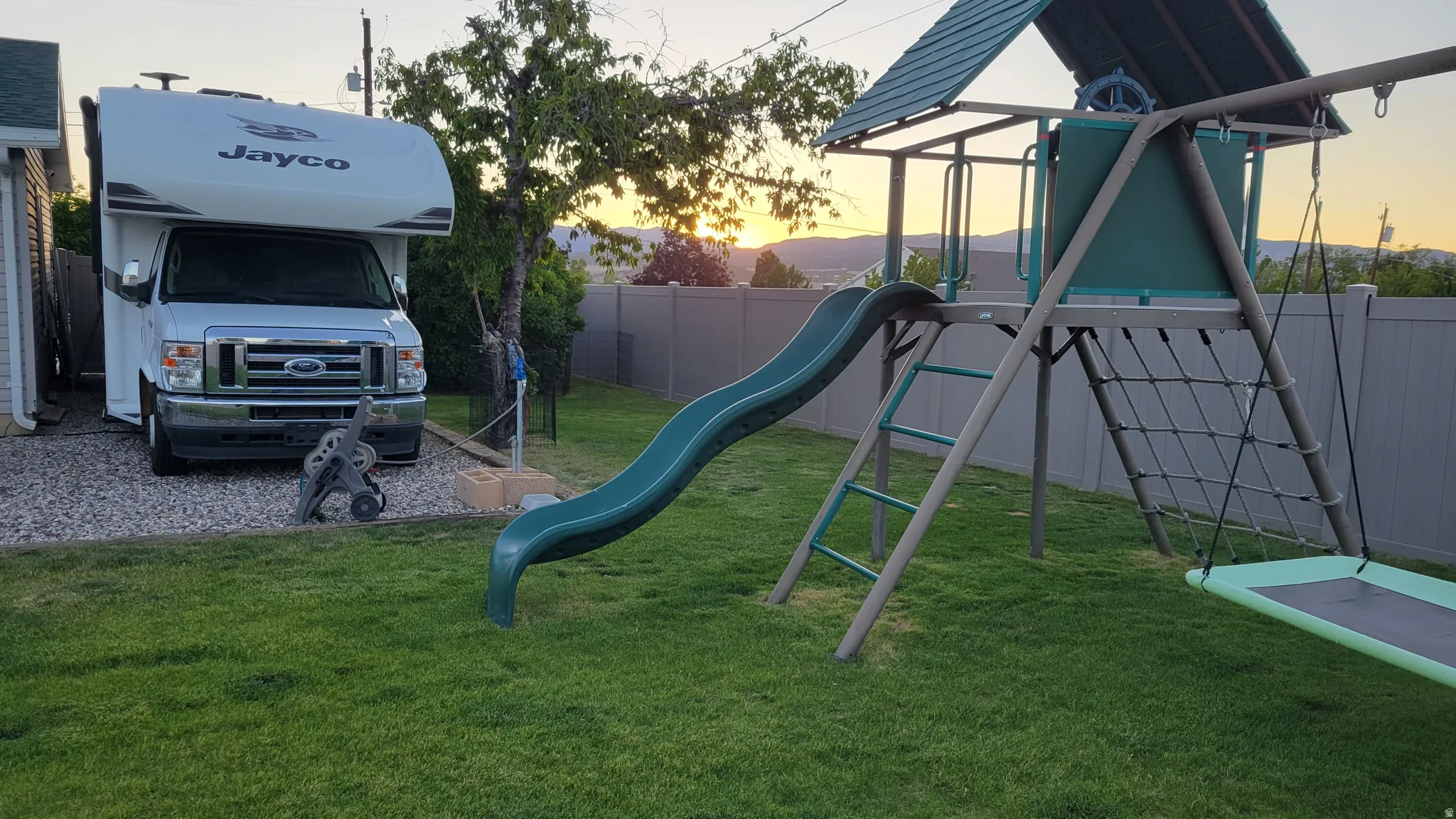Expansive backyard featuring a green lawn, a multi-component play structure with a slide and swing, a gravel parking area, a privacy fence, and a mature tree