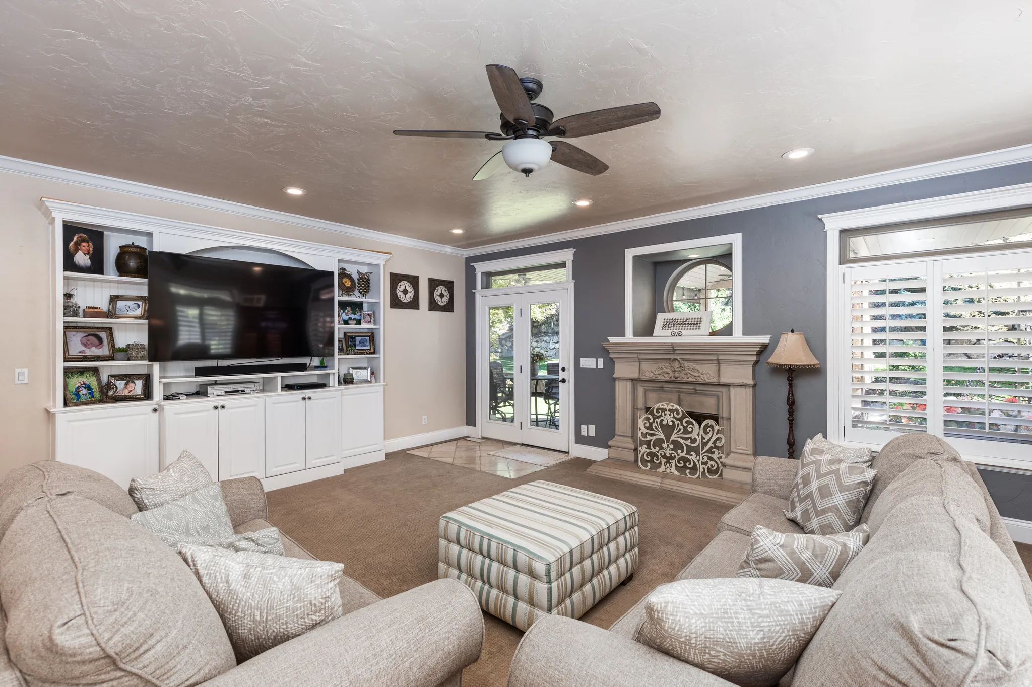 Spacious living area featuring a built-in entertainment center, a decorative fireplace with an ornate mantel, French doors, plantation shutters, and crown molding