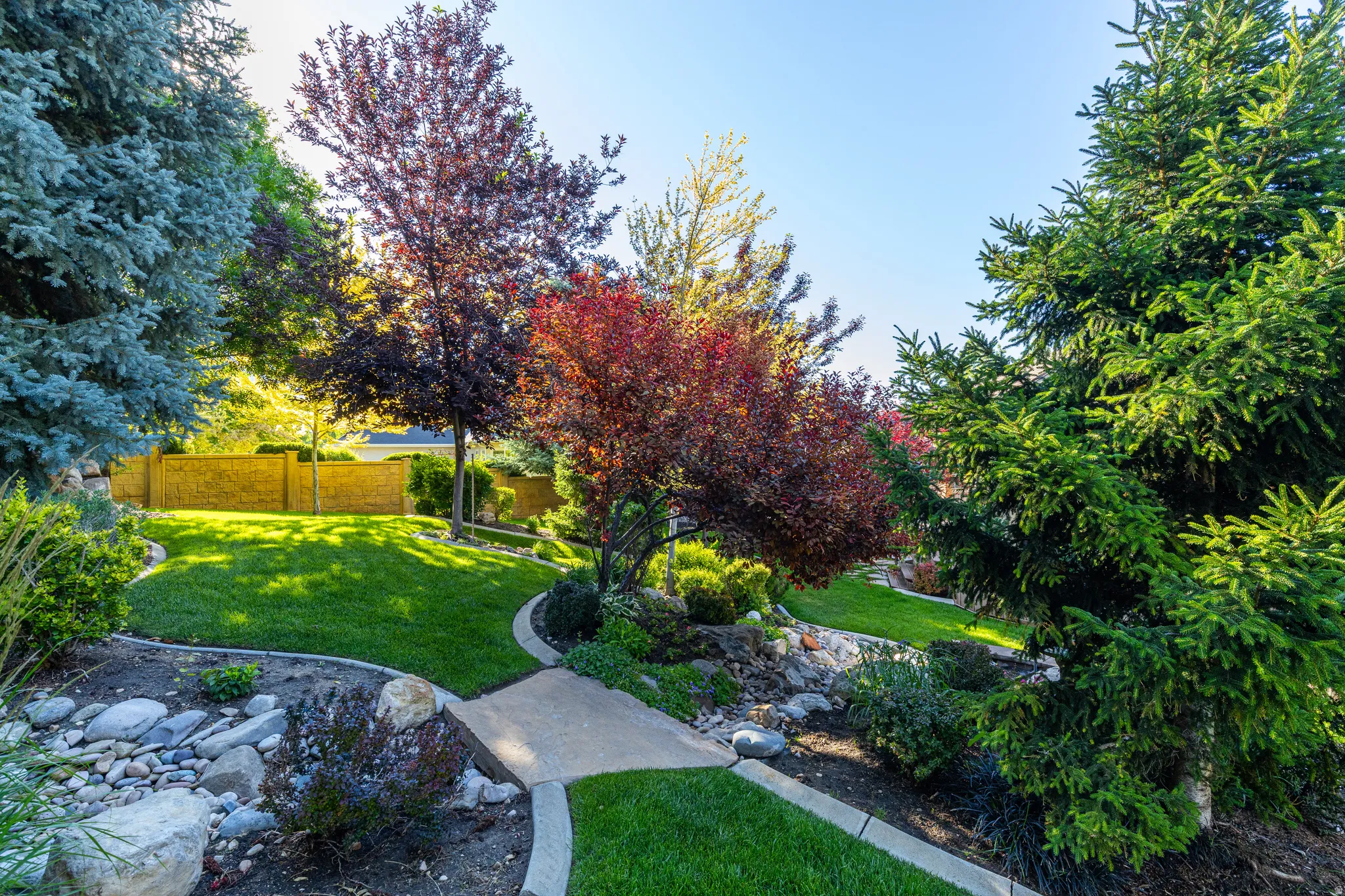 Landscaped yard featuring a curved concrete pathway, diverse mature trees, manicured green lawns, and natural rock accents