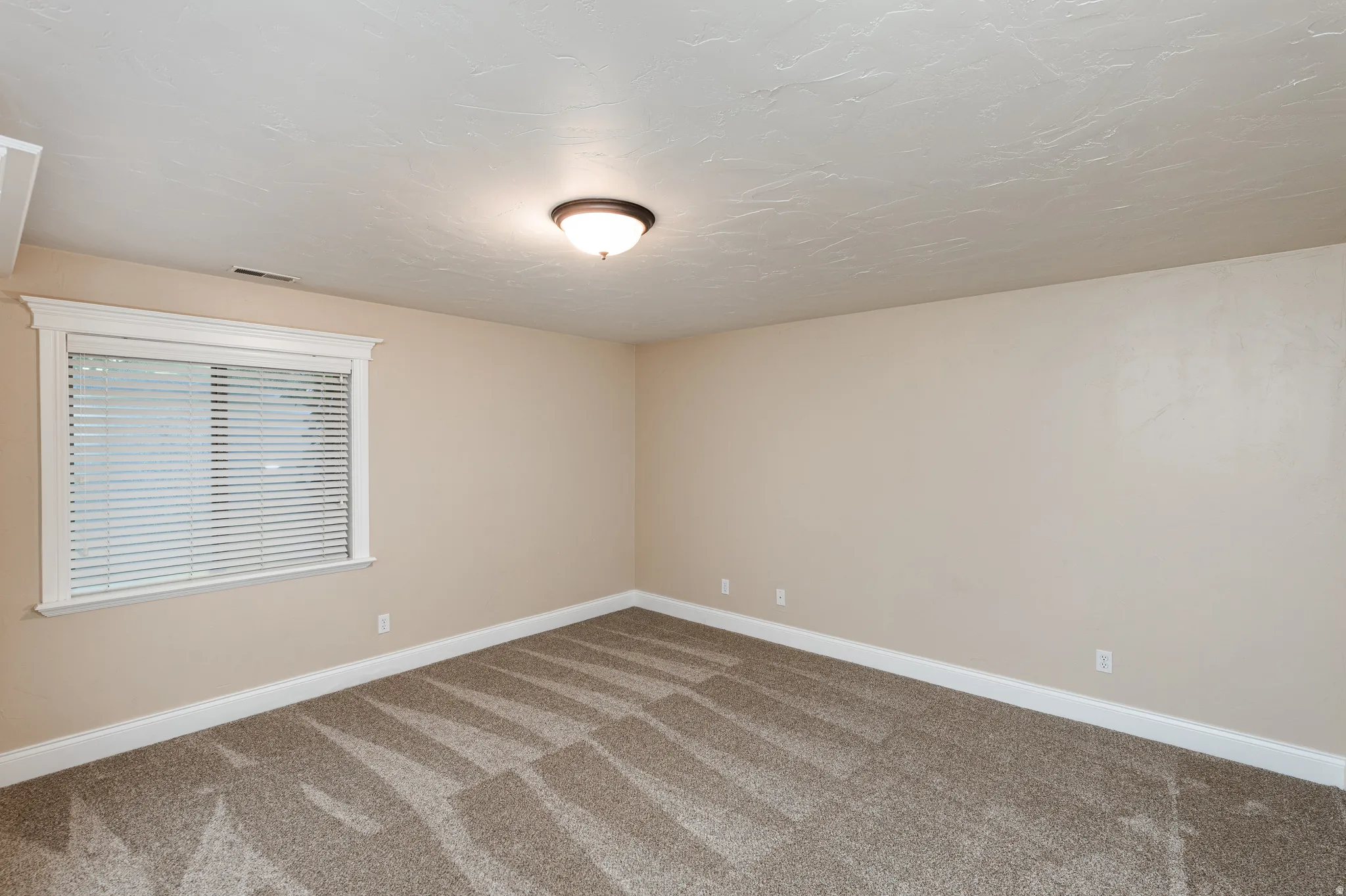 Spacious room featuring neutral paint, textured ceiling, patterned carpet, and a window with white trim and blinds