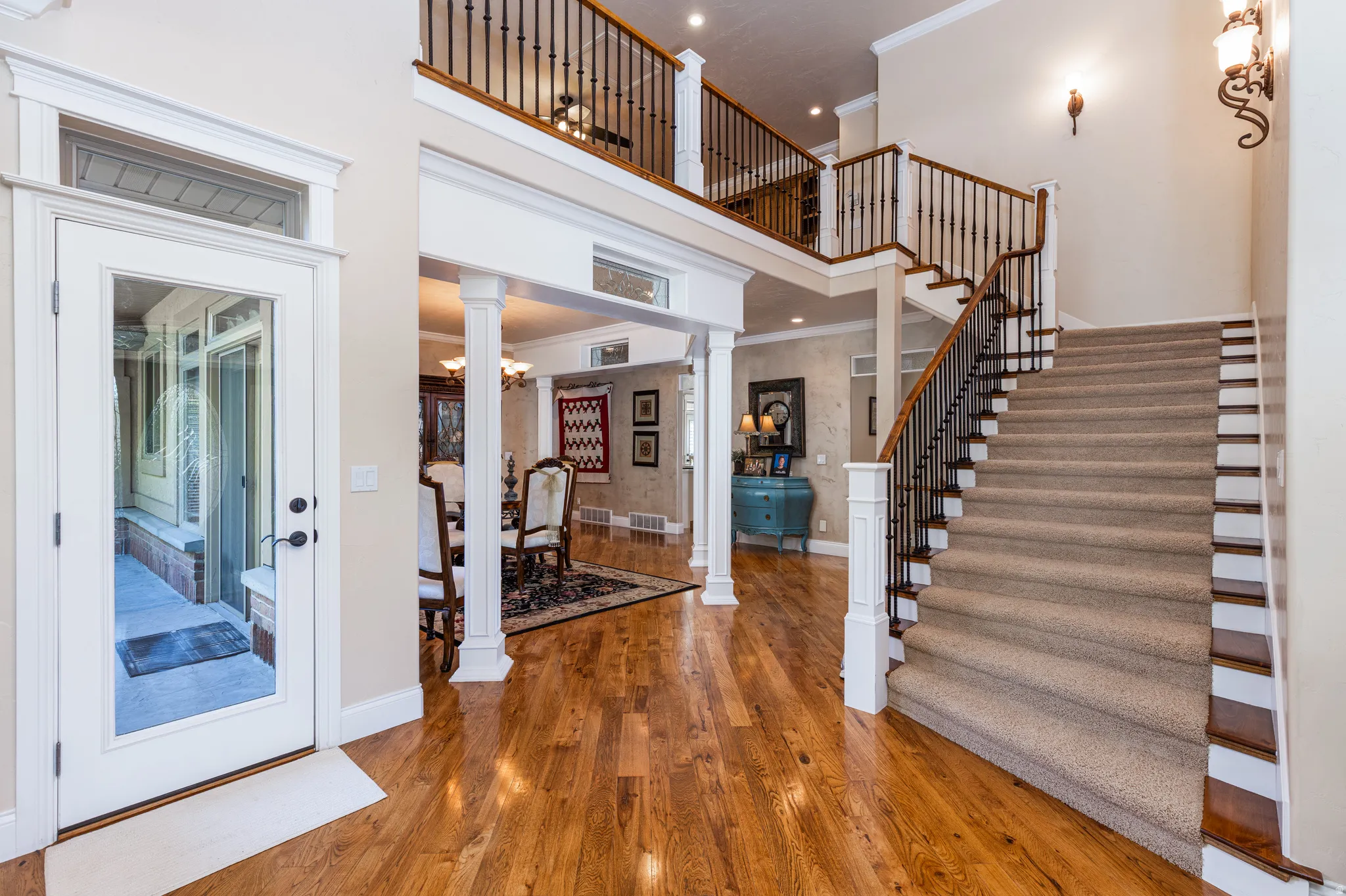 Spacious entryway featuring rich wood-finish flooring, a grand staircase with carpeted treads and wrought iron balusters, and an upper-level balcony overlooking the foyer