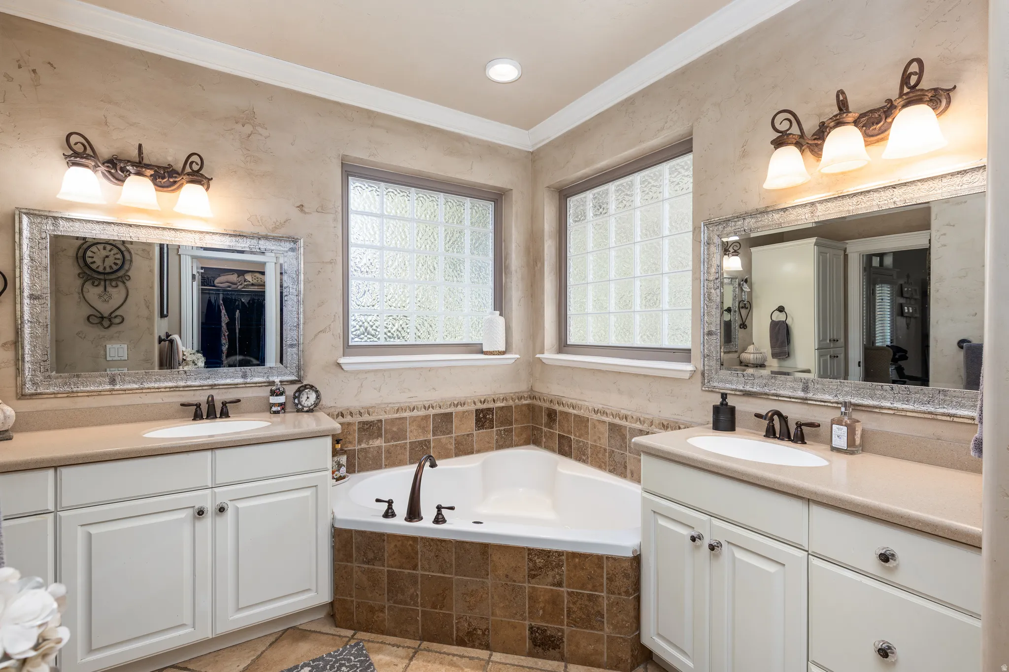 Dual vanity sinks with cream-colored countertops, an integrated corner soaking tub, textured wall finish, and two frosted glass block windows