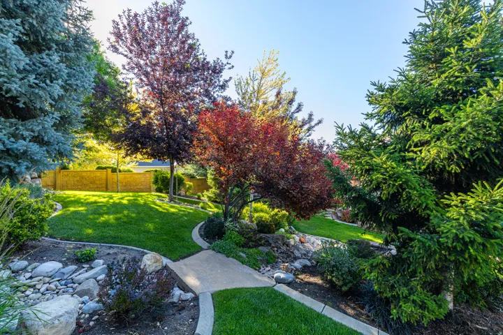 Landscaped yard featuring a curved concrete pathway, diverse mature trees, manicured green lawns, and natural rock accents