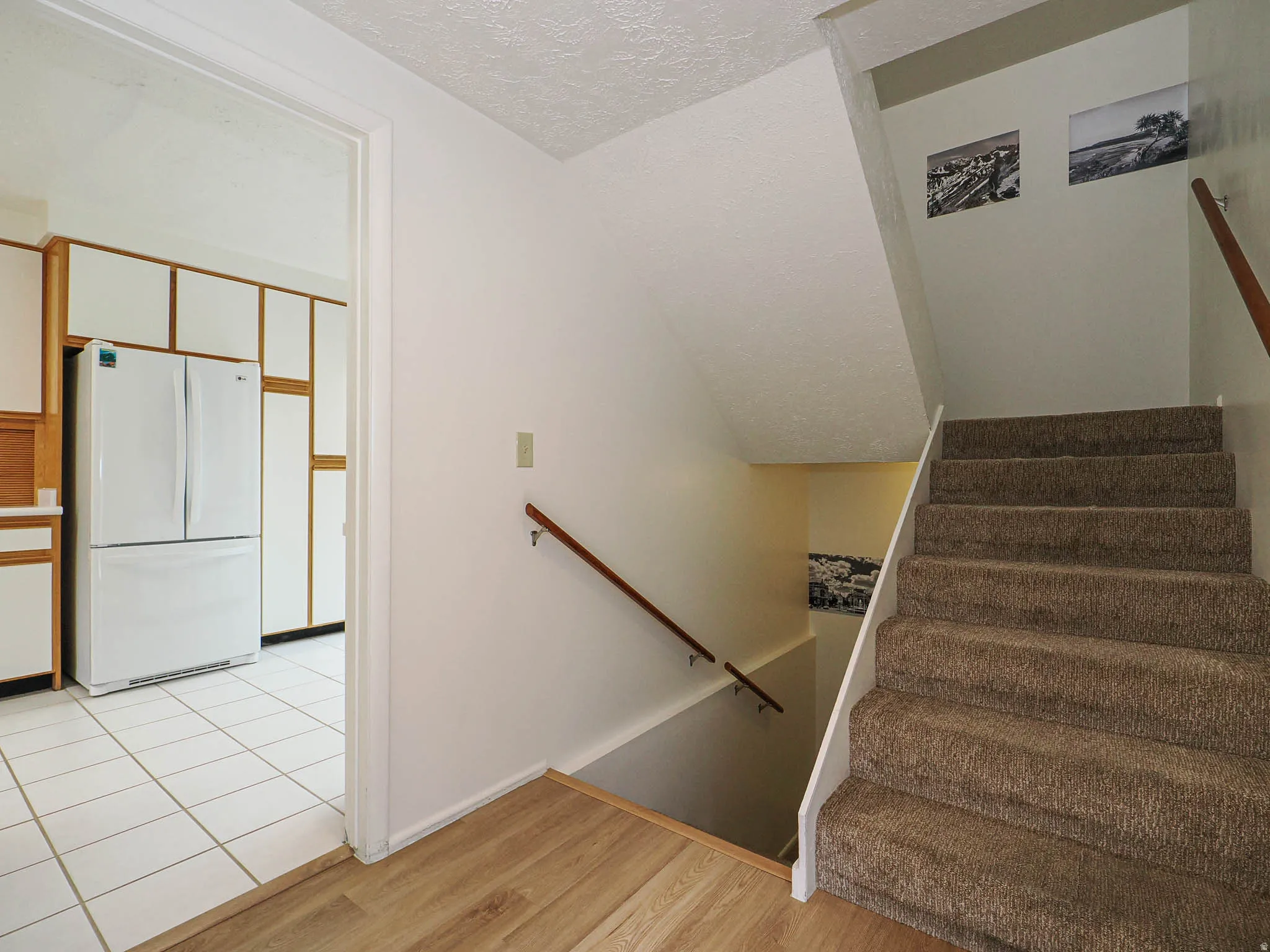 Carpeted staircase with a wood handrail, wood-finish flooring, and a kitchen featuring white appliances, wood cabinetry, and white tile flooring