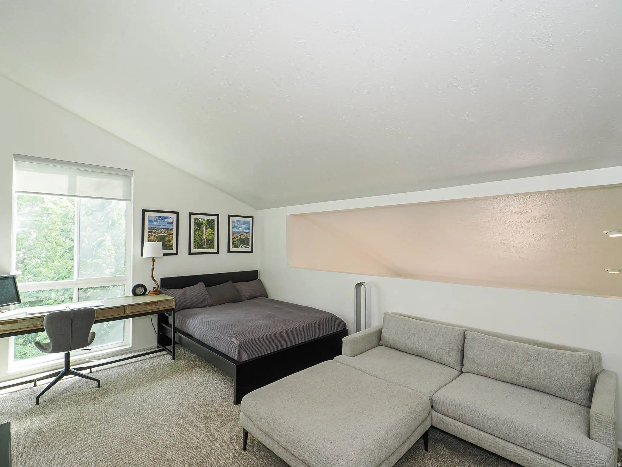 Room featuring an angled ceiling, large window with roller shade, and a light-colored carpeted floor