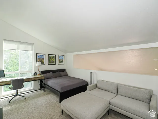 Room featuring an angled ceiling, large window with roller shade, and a light-colored carpeted floor