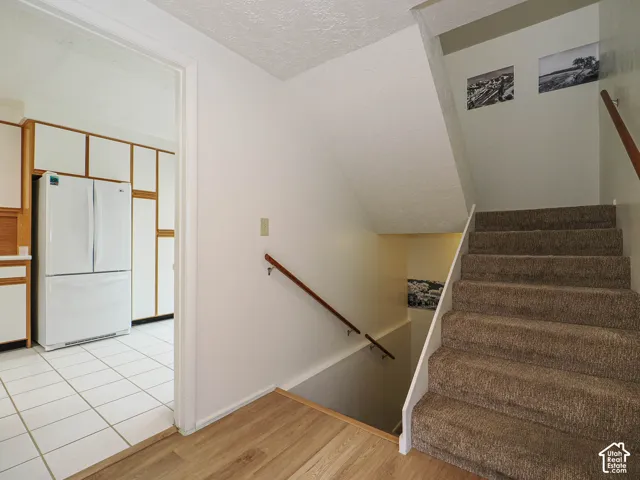 Carpeted staircase with a wood handrail, wood-finish flooring, and a kitchen featuring white appliances, wood cabinetry, and white tile flooring