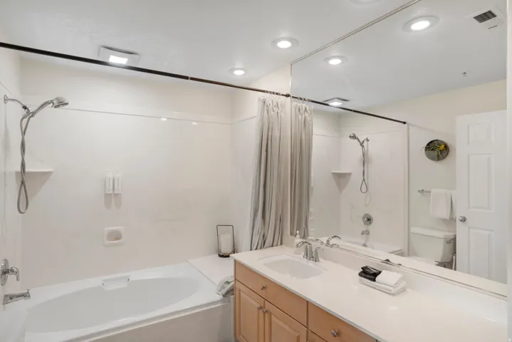 Bathroom featuring a soaking tub with an integrated shower, a vanity with a light-toned countertop, a single basin sink, wood-finish cabinetry, and a full-width wall mirror