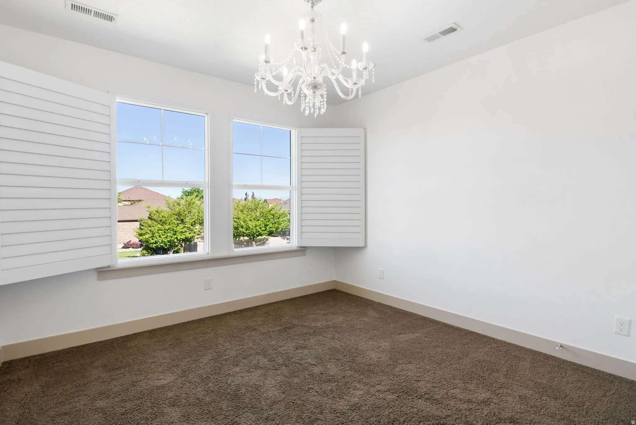 Room featuring a decorative chandelier, dual windows with white plantation shutters, white walls, and brown carpet flooring