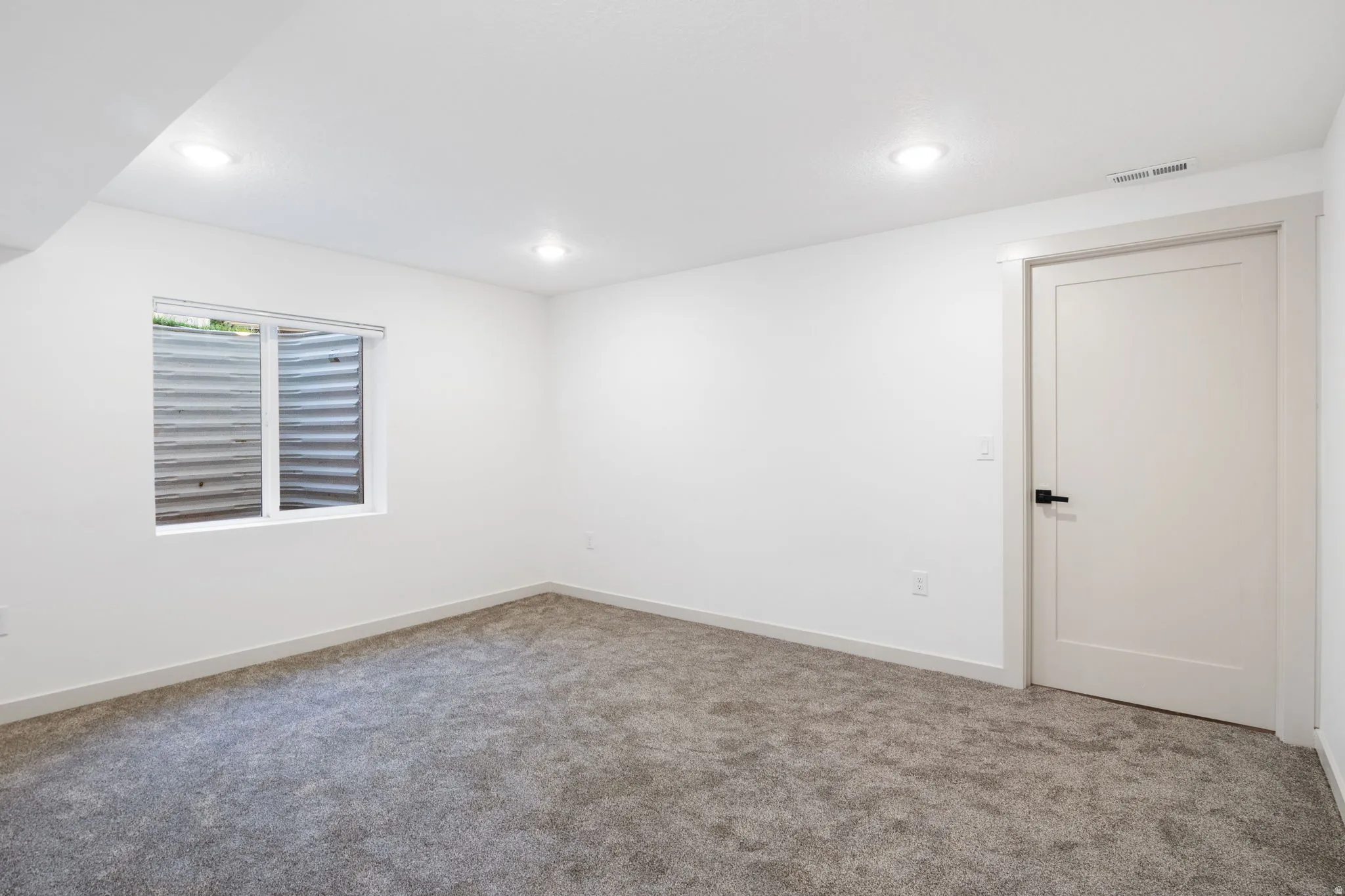 Carpeted room featuring a white interior door with matte black hardware, a window with a window well, recessed lighting, and white baseboards