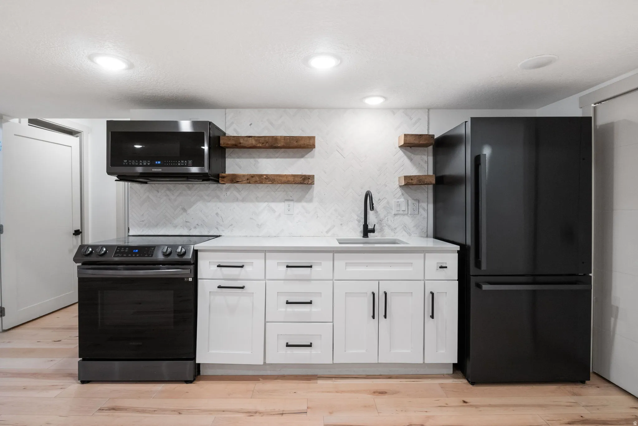 Modern kitchen featuring black stainless steel appliances, white cabinetry with matte black hardware, light wood-finish flooring, and a herringbone pattern tile backsplash