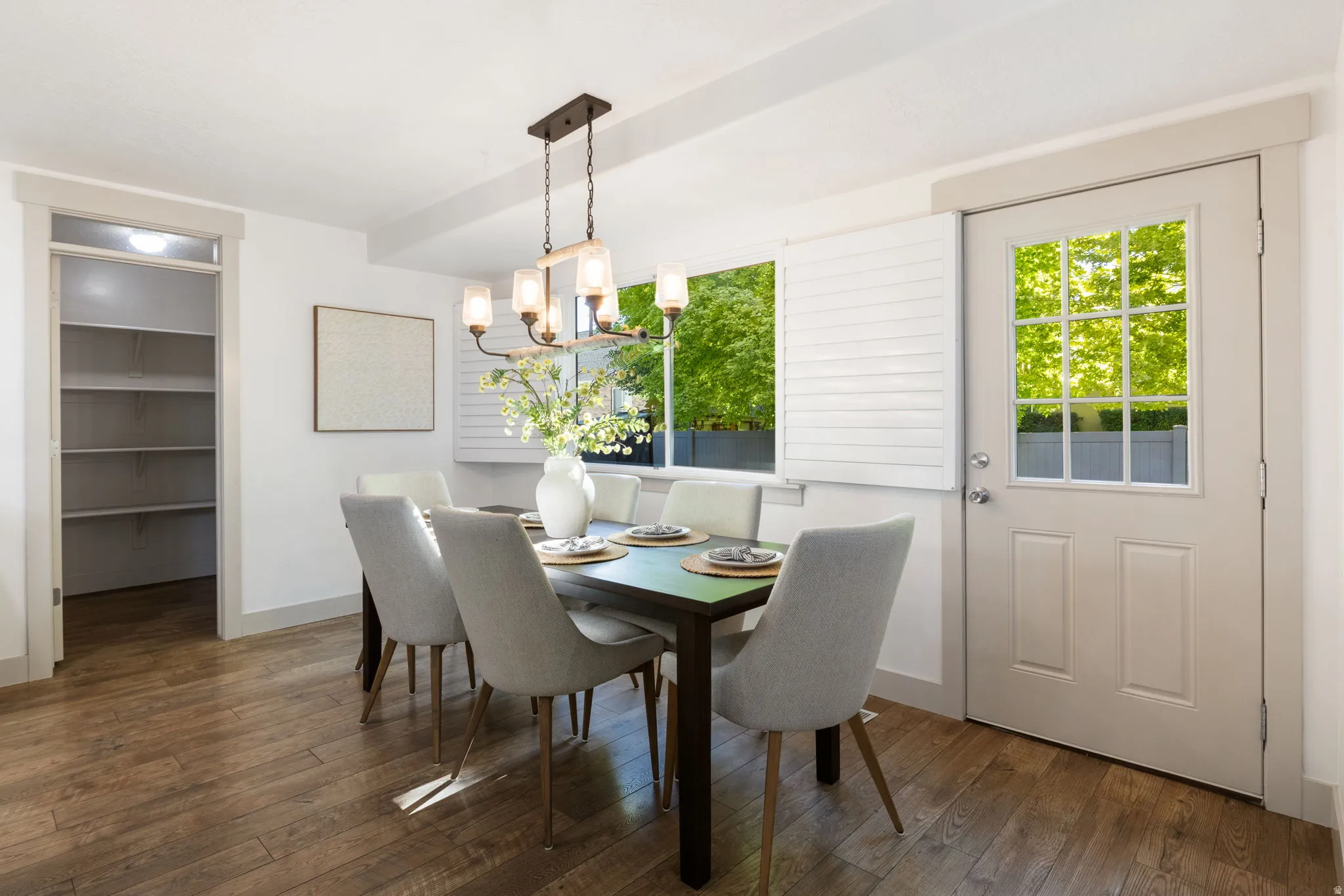 Dining area featuring wood-finish flooring, a contemporary chandelier, two windows with white plantation shutters, and a paneled door with glass inserts