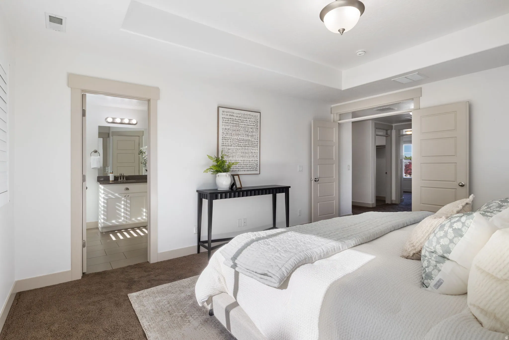 Spacious bedroom featuring neutral carpeting, a tray ceiling with a semi-flush mount light fixture, and a doorway to a bathroom with a vanity and stone-finish countertop