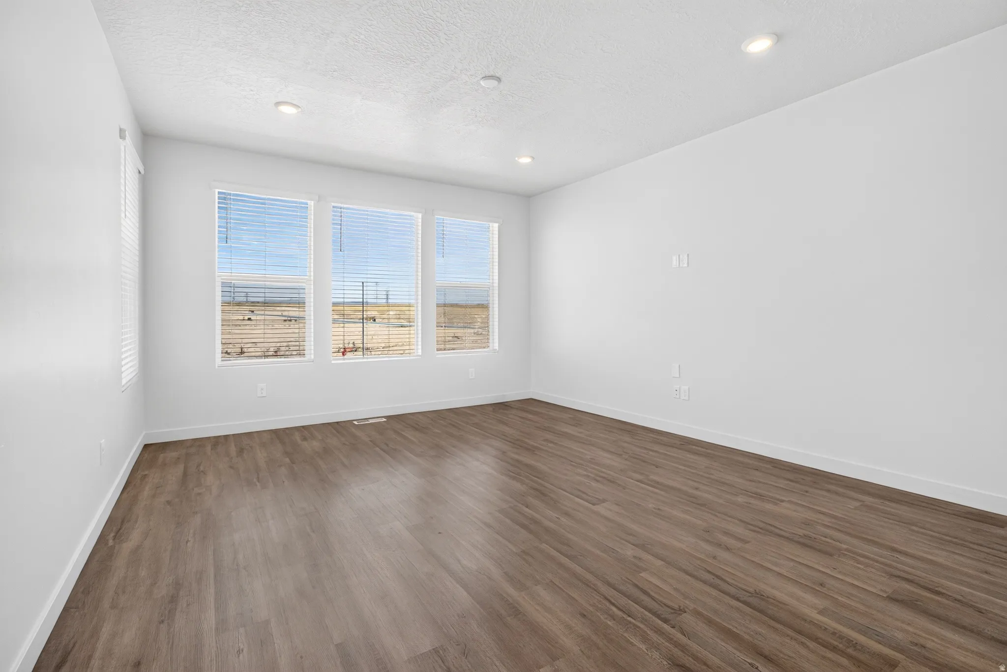 Spacious room featuring three large windows with horizontal blinds, wood-finish flooring, white baseboards, recessed ceiling lighting, and a neutral color palette