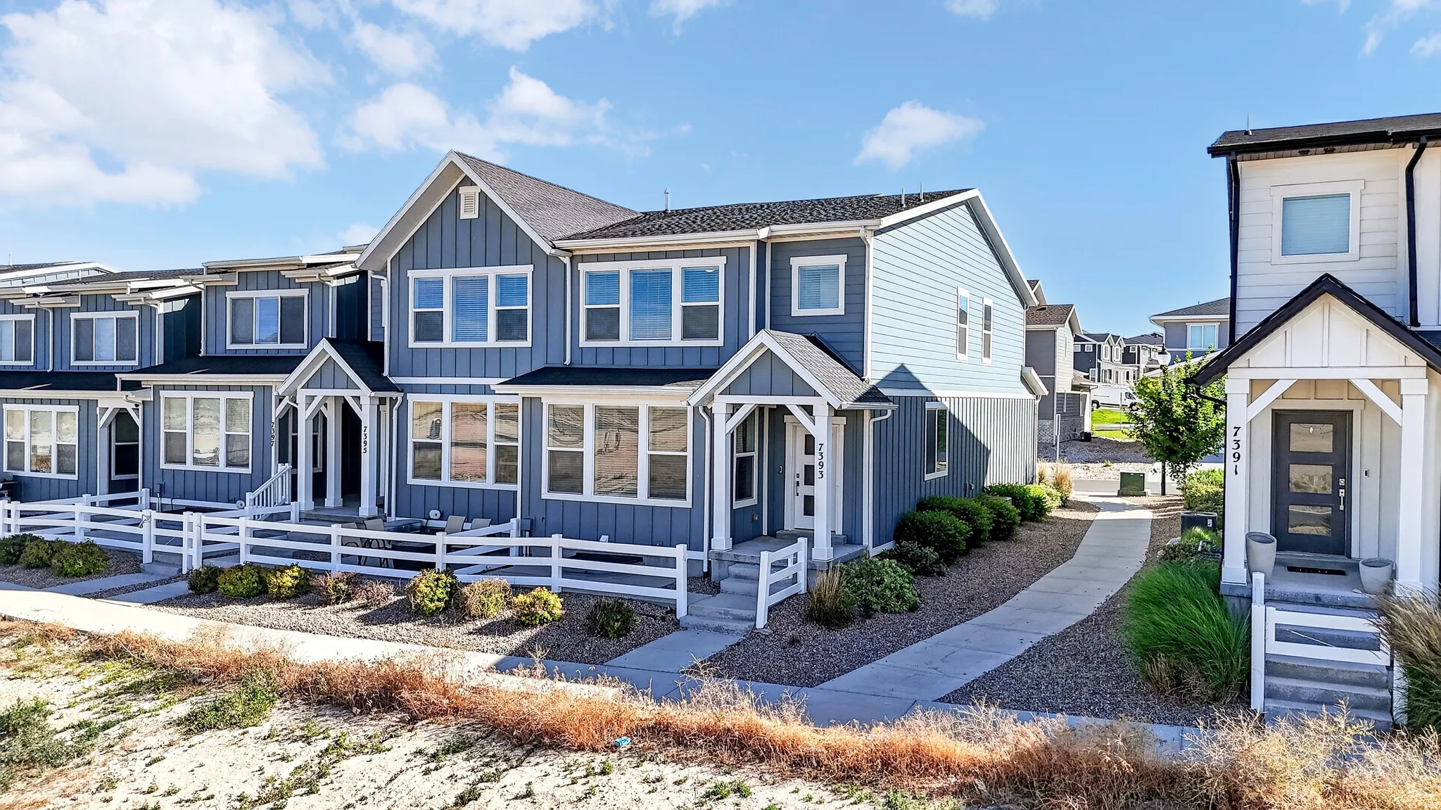 Modern townhome facade featuring a blue vertical siding, white trim, gabled entryways, and white picket fencing