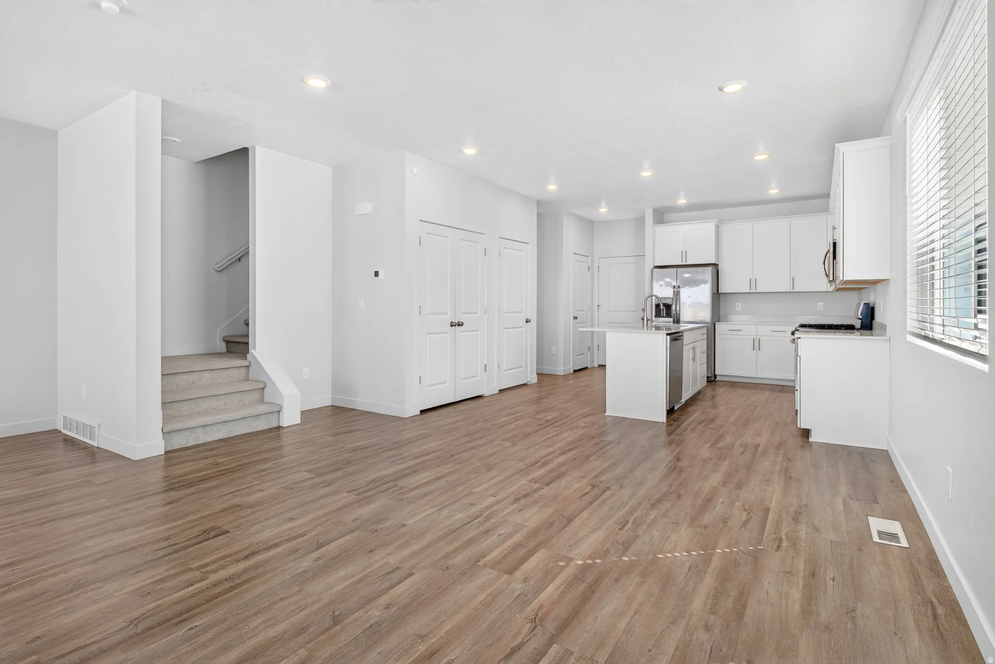 Open-concept living area featuring wood-finish flooring, a contemporary kitchen with white cabinetry, stainless steel appliances, and a central island with a sink