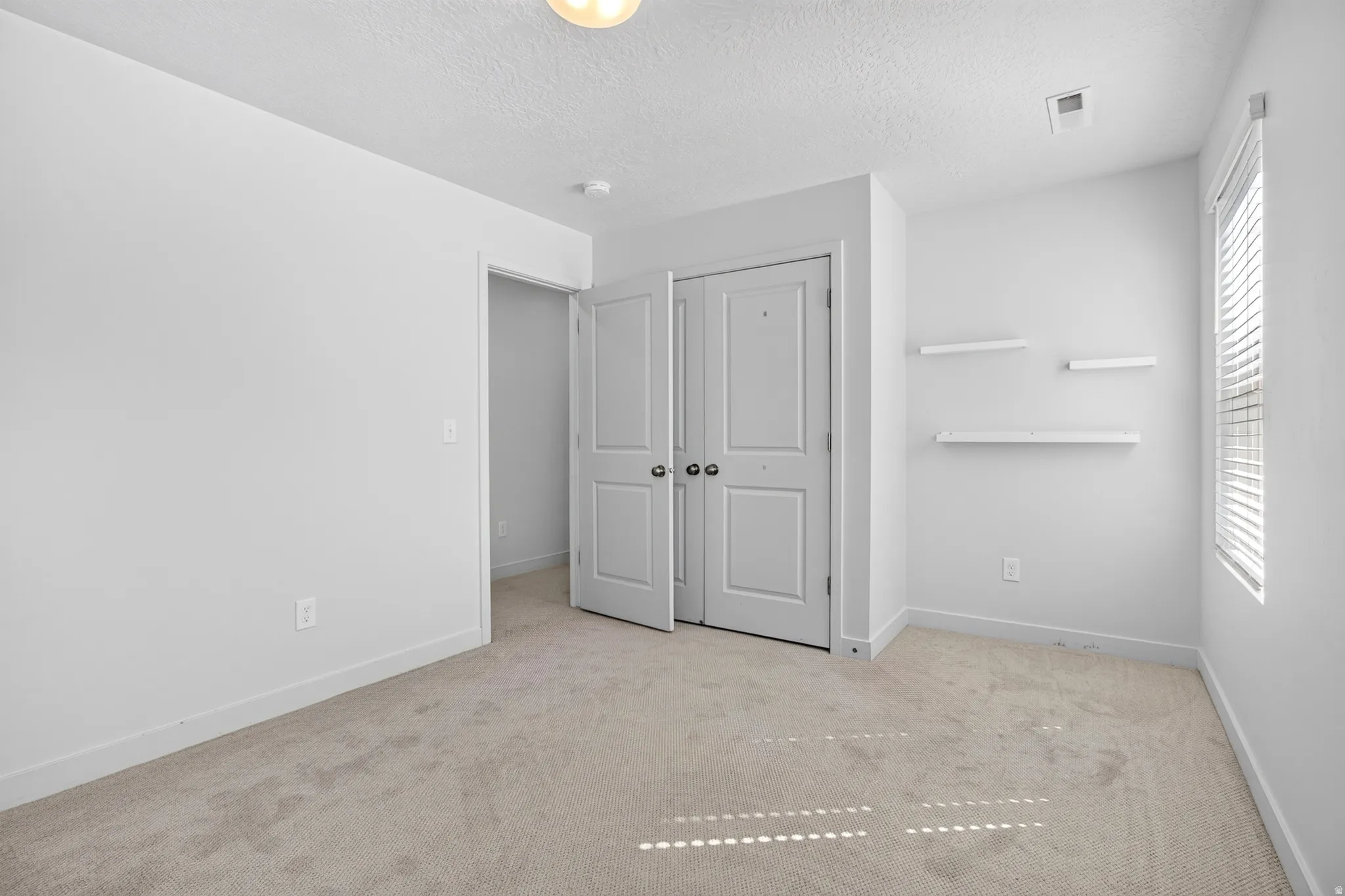 Neutral tone wall paint, light-colored carpeting, white baseboards, and a window with horizontal blinds