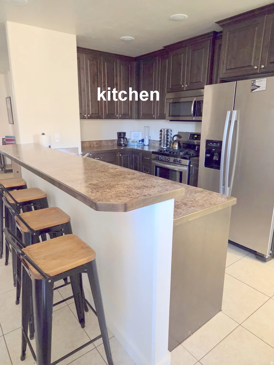 Open-concept kitchen featuring dark wood-finish cabinetry, stainless steel appliances, speckled countertops, and a breakfast bar with seating