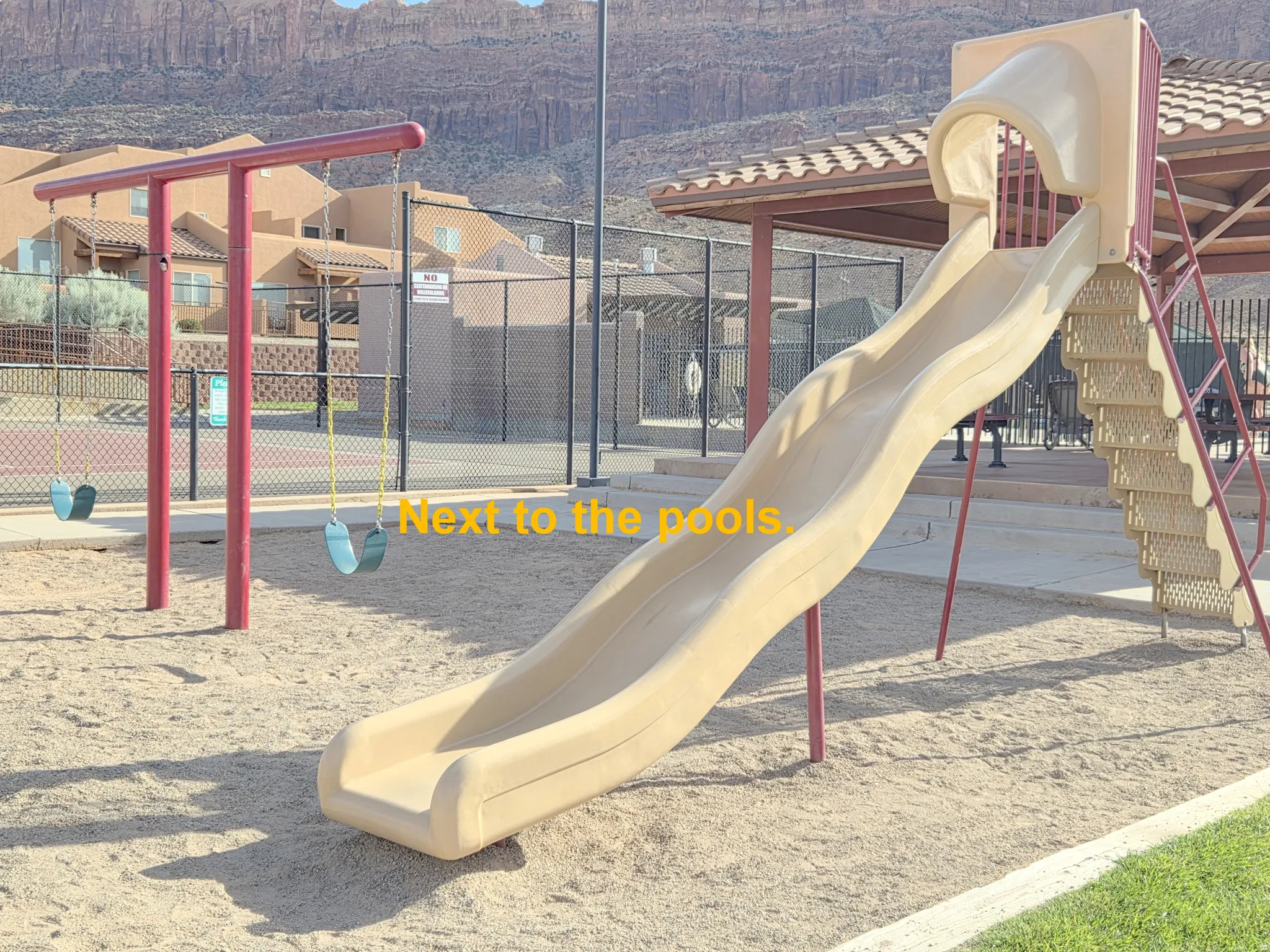 Community playground featuring a sand surface, a beige slide with ladder access, and a red-framed swing set with two swings