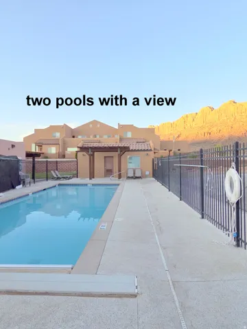 Two swimming pools featuring a surrounding concrete deck, black metal fencing, and a nearby building with a covered patio area, all set against a backdrop of natural rock formations