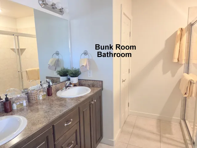 Bunk Room Bathroom featuring a dual vanity with a patterned countertop and white oval sinks, dark wood-finish cabinetry, a large wall-mounted mirror, and a shower with a sliding glass door and tiled walls