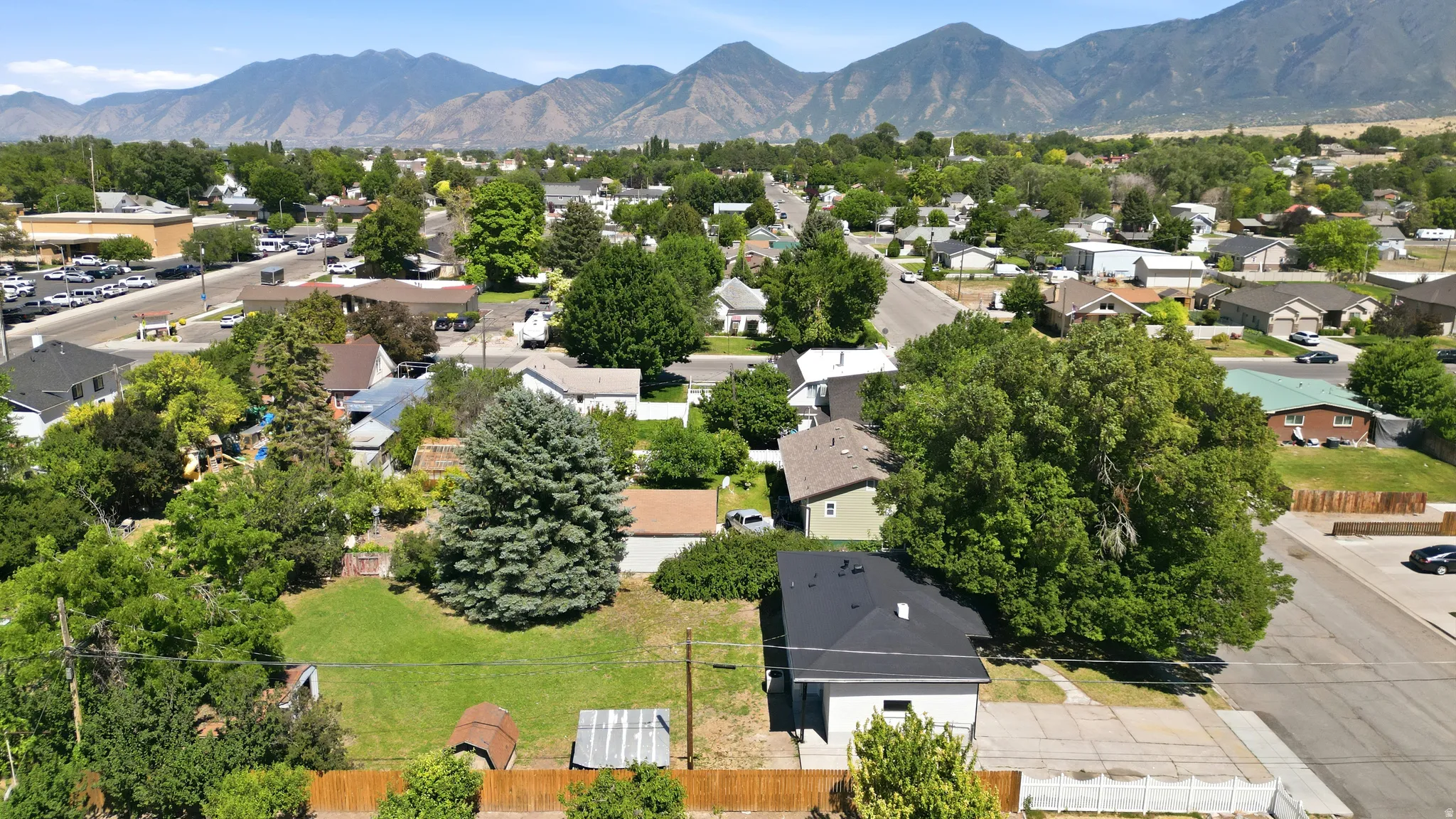 Aerial perspective showcasing the property with a dark roof and a paved driveway, surrounded by mature trees and a green lawn