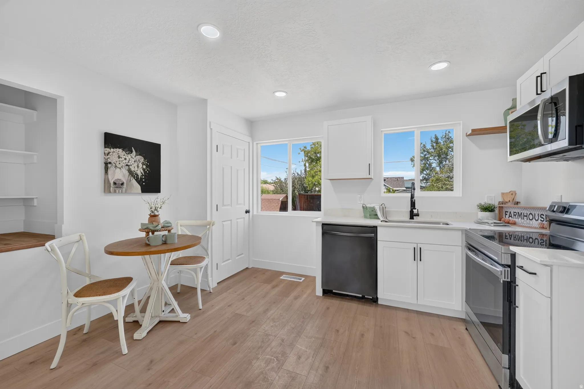 Contemporary kitchen featuring white cabinetry, light-toned wood-finish flooring, two large windows, recessed ceiling lighting, and a suite of stainless steel appliances