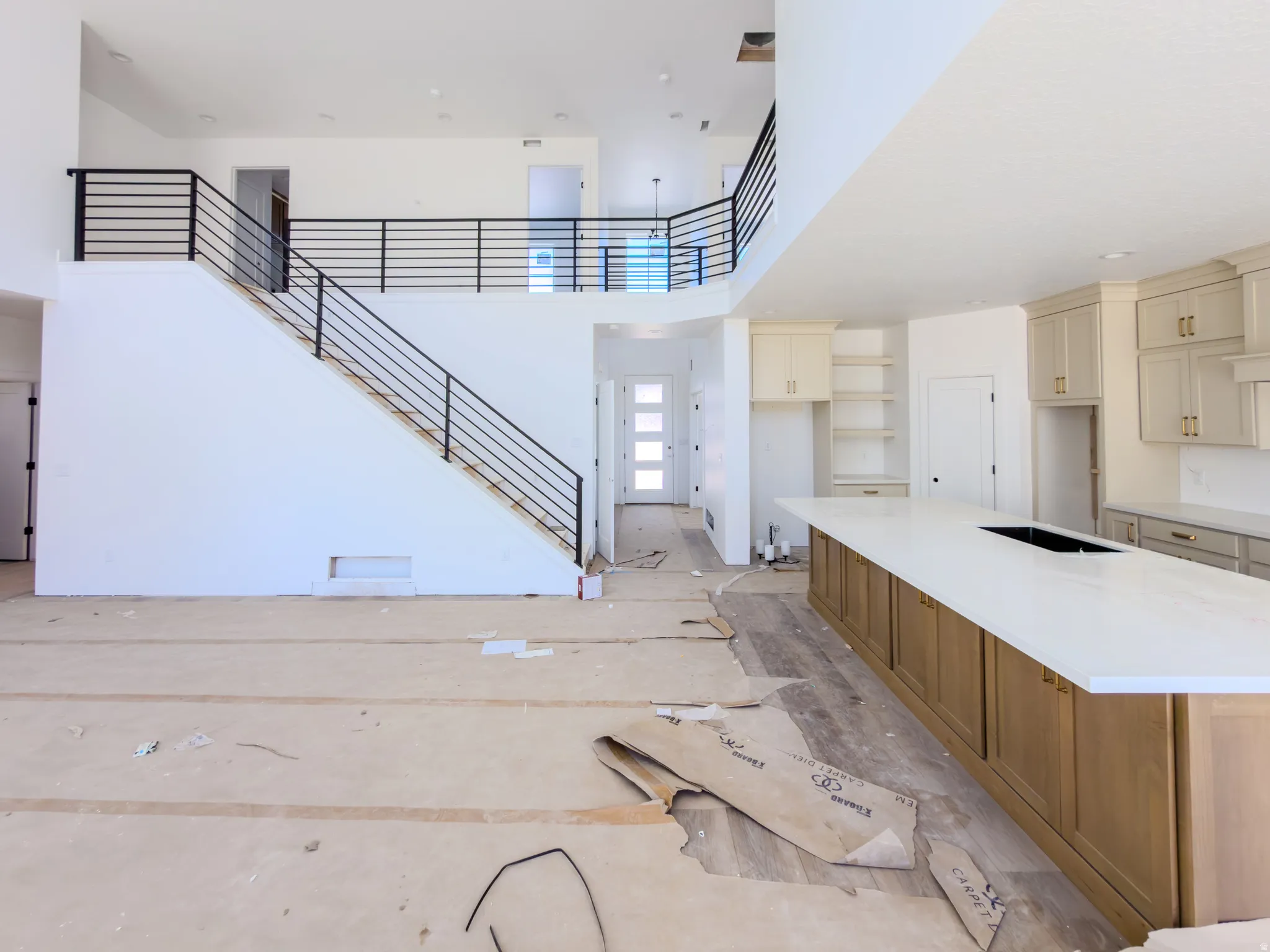 Open-concept living space featuring a two-story ceiling, modern iron stair railing, a large kitchen island with a white countertop, light-toned wood-finish cabinetry, and neutral wall paint