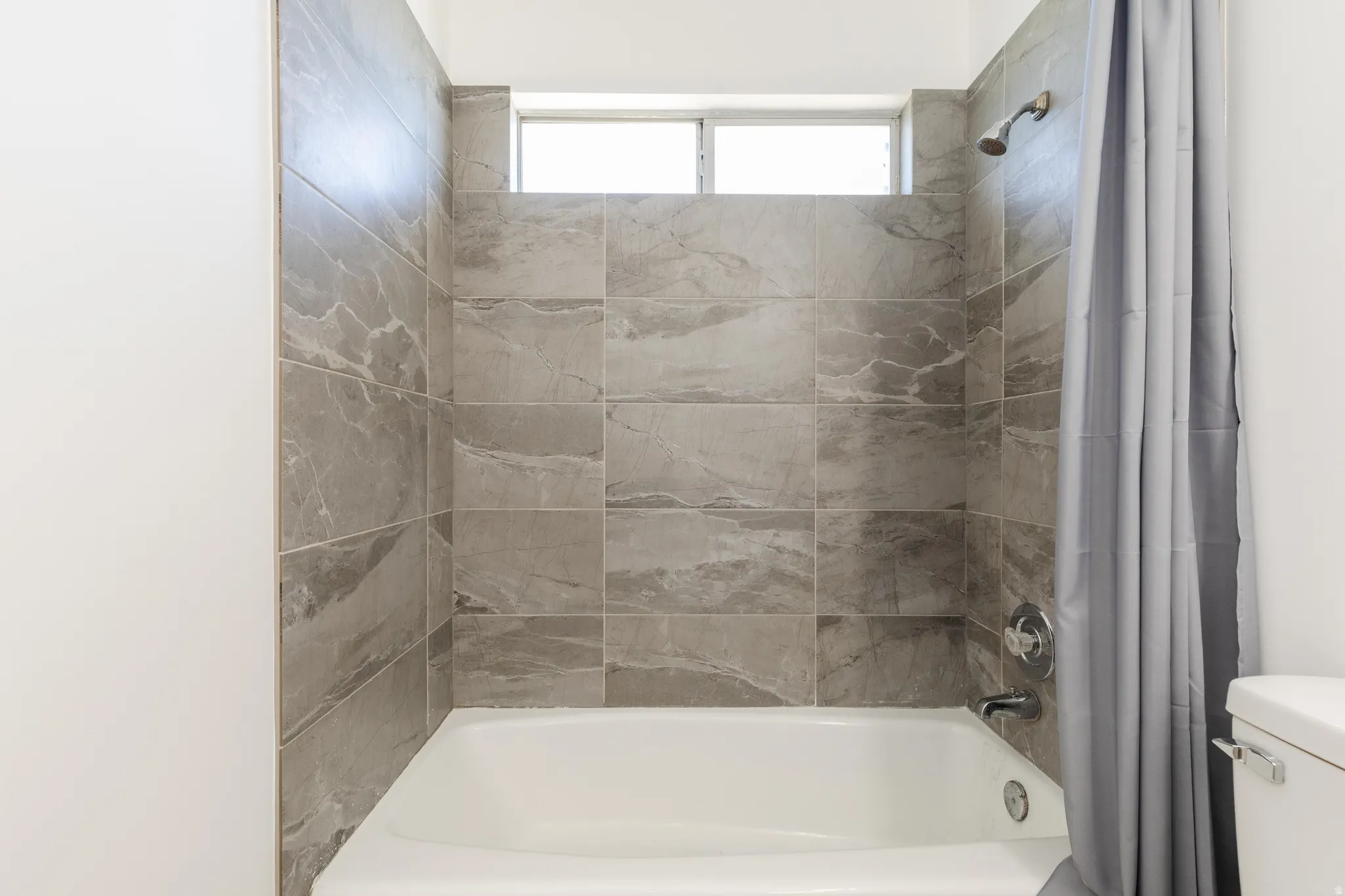 Bathtub with a tiled surround featuring large format gray stone-look tiles, a chrome showerhead, and a small transom window