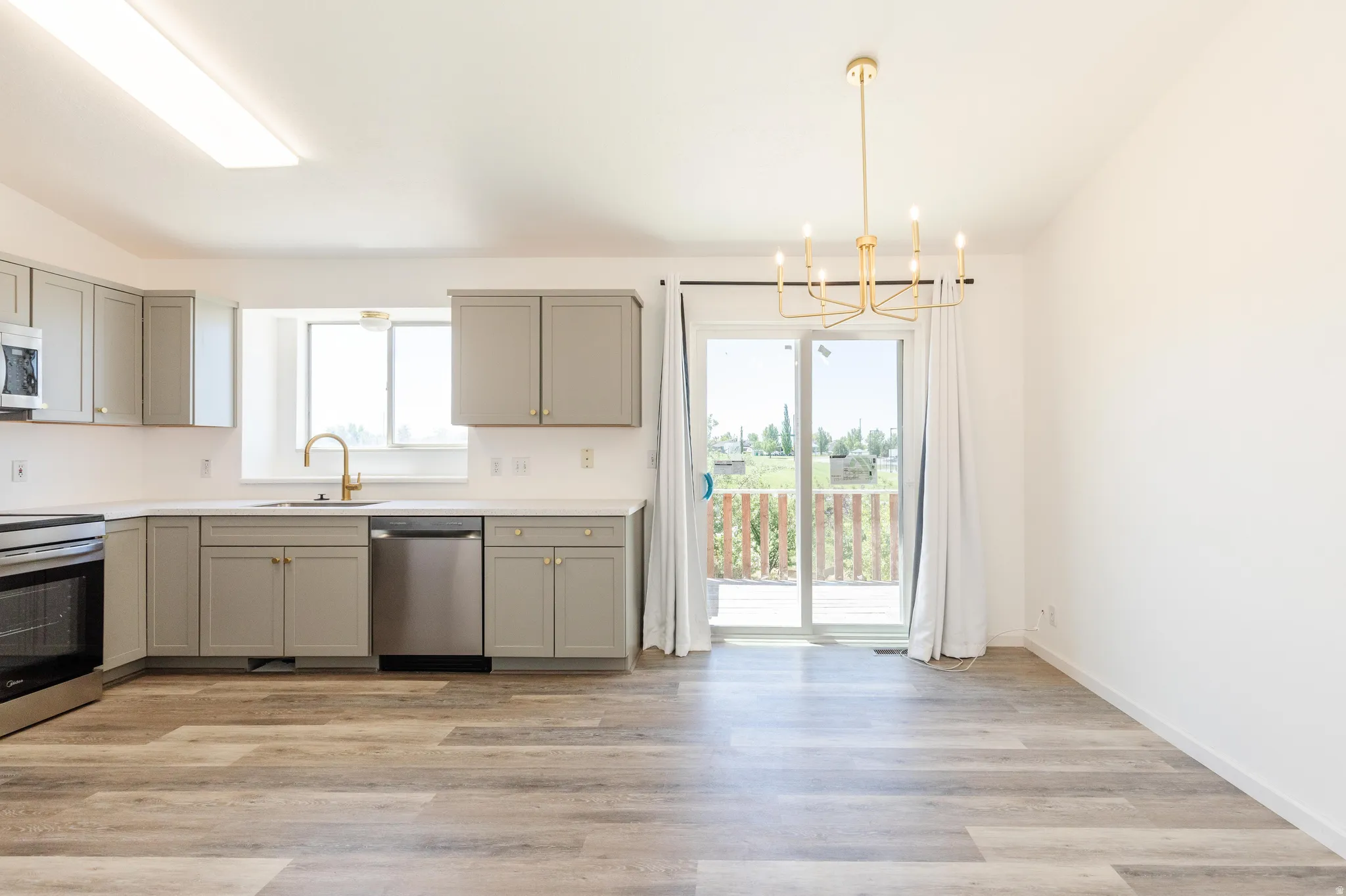 Open-concept area featuring wood-finish flooring, a modern gold-tone chandelier, and a kitchen with gray cabinetry, white countertops, and stainless steel appliances