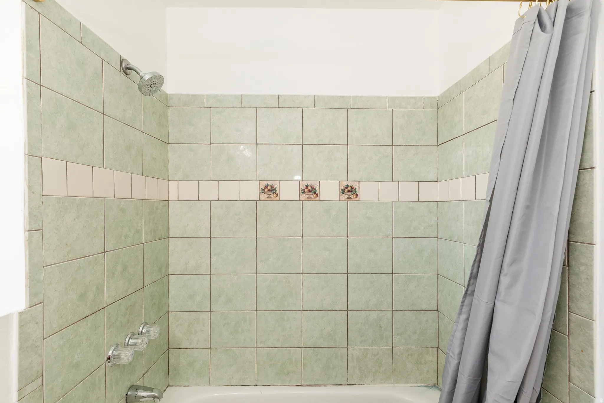 Bathtub with light green tiled surround featuring a decorative accent band and chrome shower fixtures