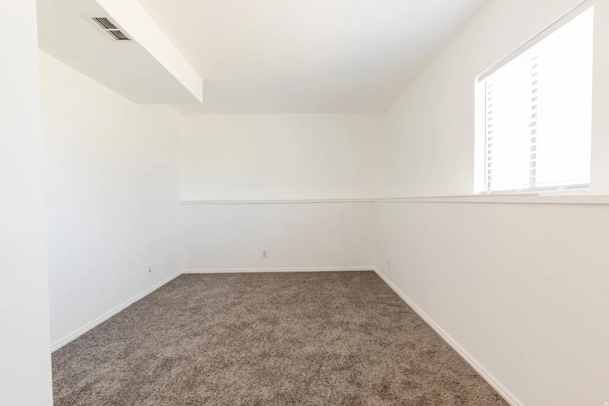 Spacious room featuring neutral carpeting, white painted walls, and a window with horizontal blinds