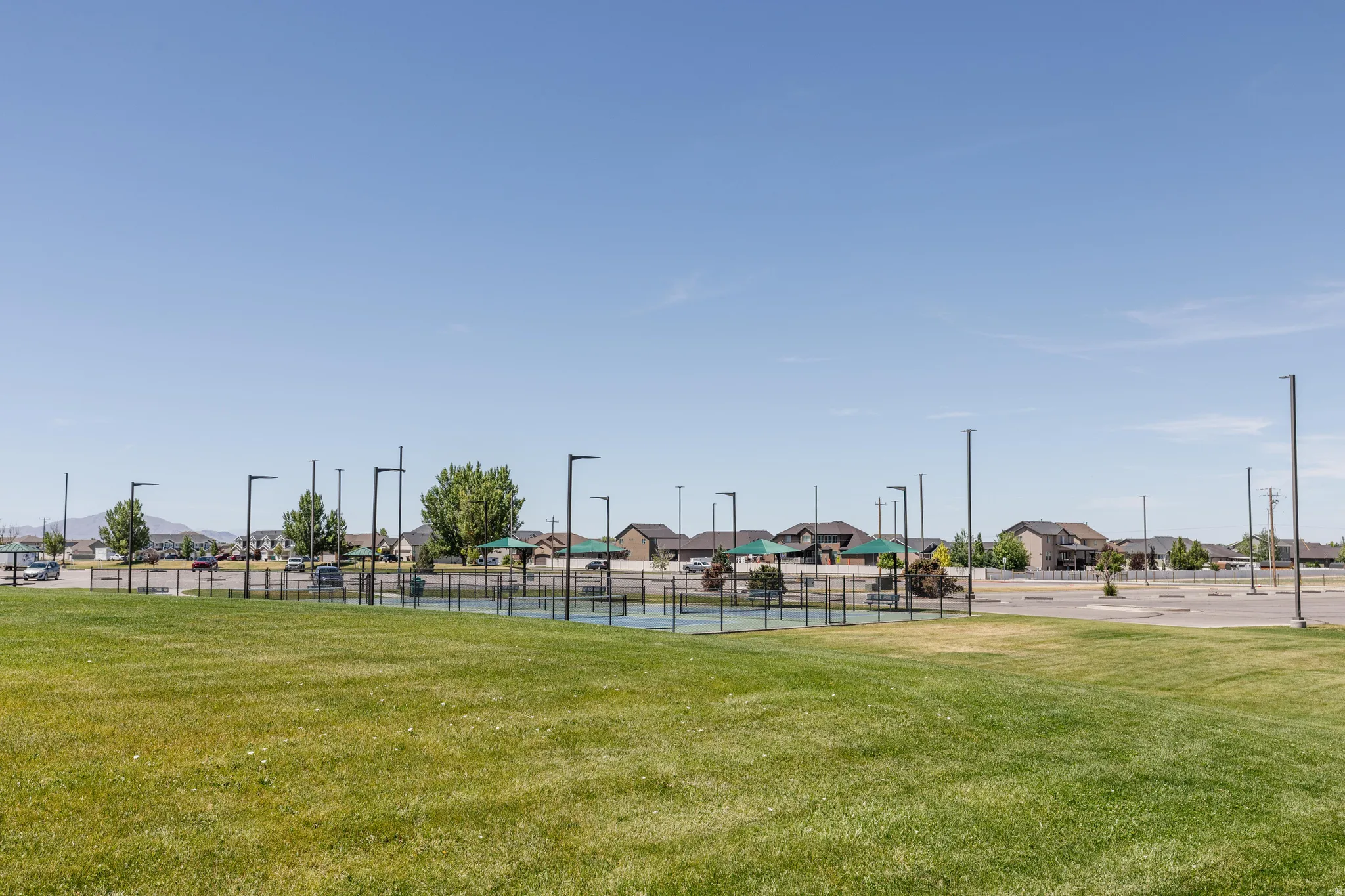 Expansive green space featuring a hard-surface court with fencing, surrounded by numerous light poles, and a paved parking area