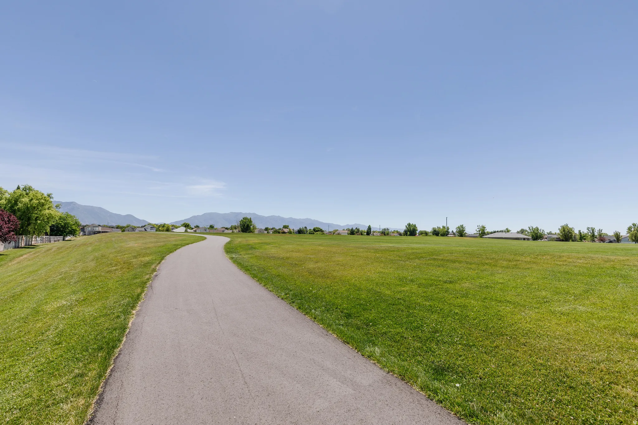Paved walking path bordered by expansive green space, featuring distant mountain ranges and suburban architecture