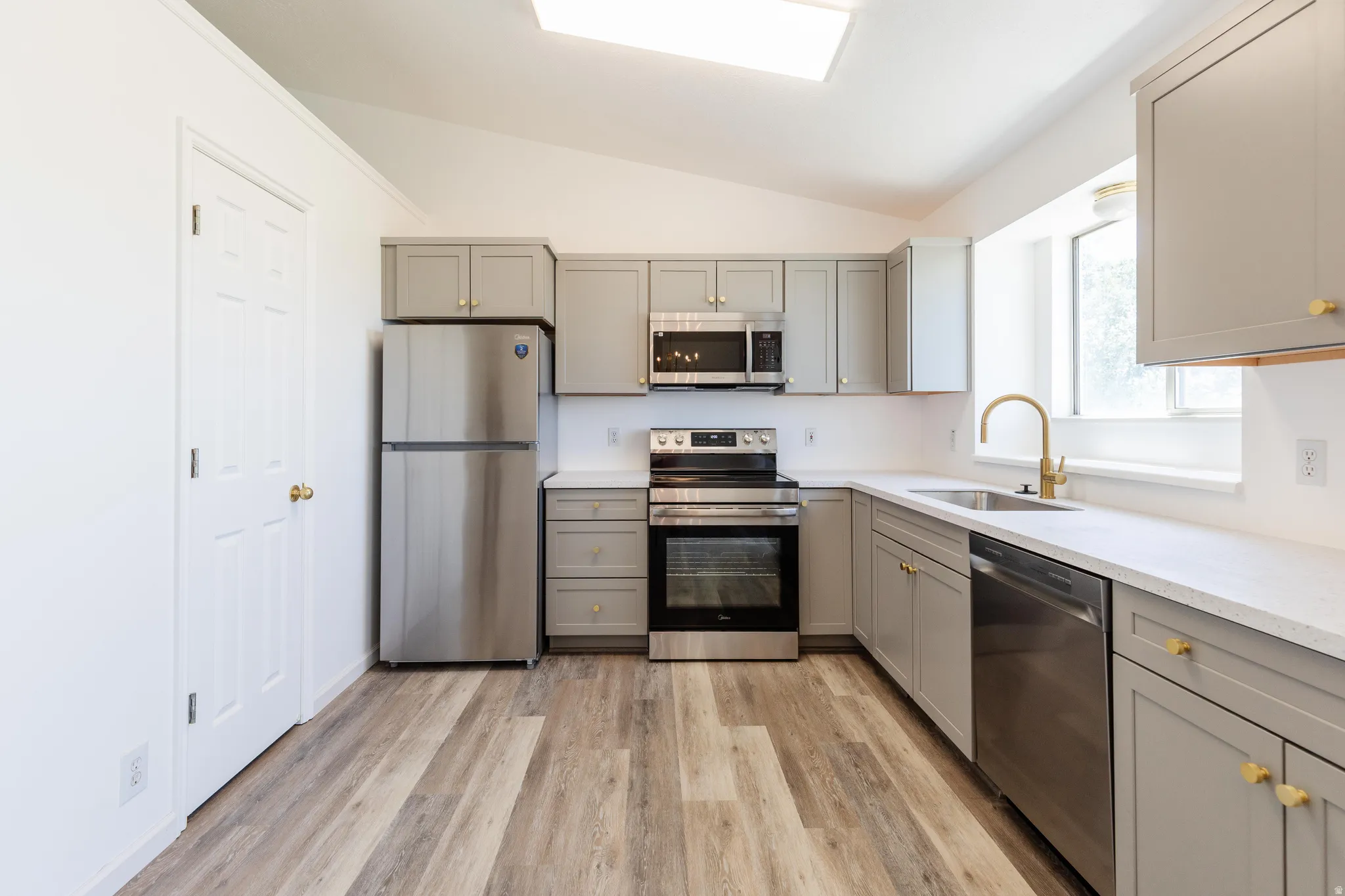 Contemporary kitchen featuring light gray cabinetry with gold-tone hardware, stainless steel appliances, wood-finish flooring, white countertops, and a gold-tone gooseneck faucet