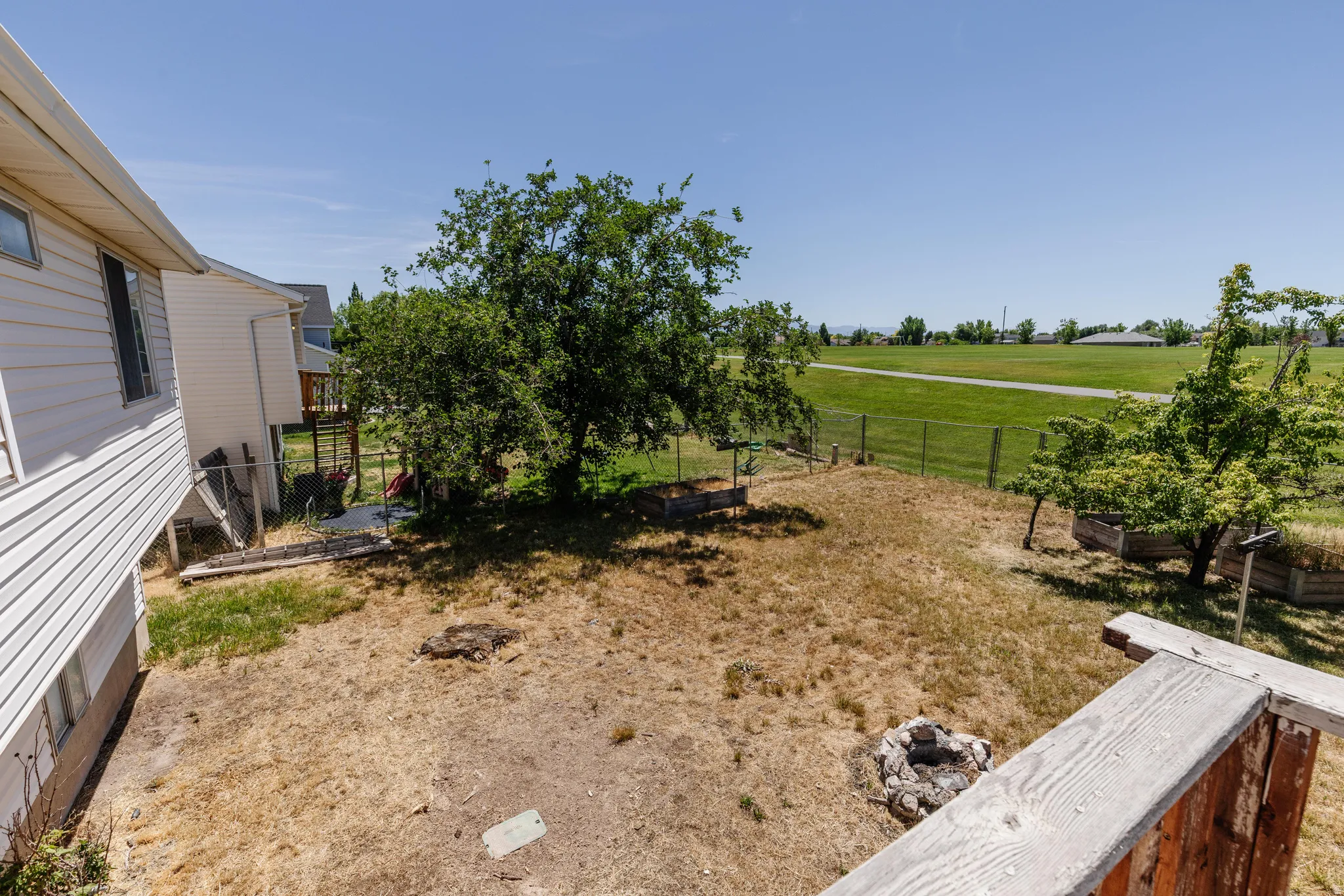 Expansive backyard featuring mature trees, raised garden beds, a built-in fire pit, and a wire fence bordering an open field