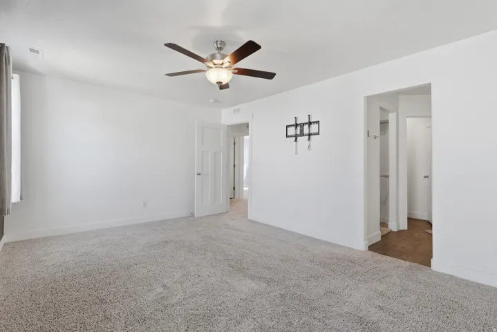 Spacious room featuring light gray carpeting, crisp white walls, a ceiling fan with integrated lighting, and a window with gray drapes