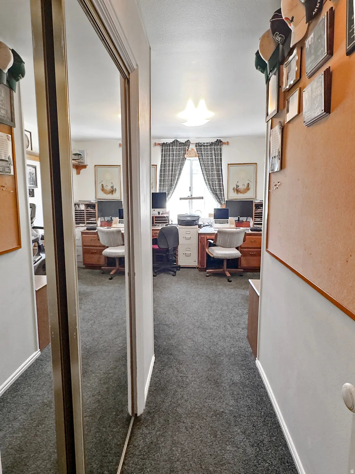 Dedicated office space featuring two built-in desk areas with wood cabinetry, a central window with patterned drapes, a flush-mount ceiling light fixture, and dark gray carpet flooring