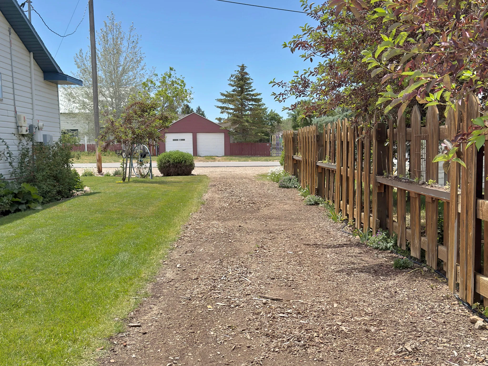 Unpaved driveway running alongside a wooden picket fence with horizontal plank accents, bordered by a manicured lawn