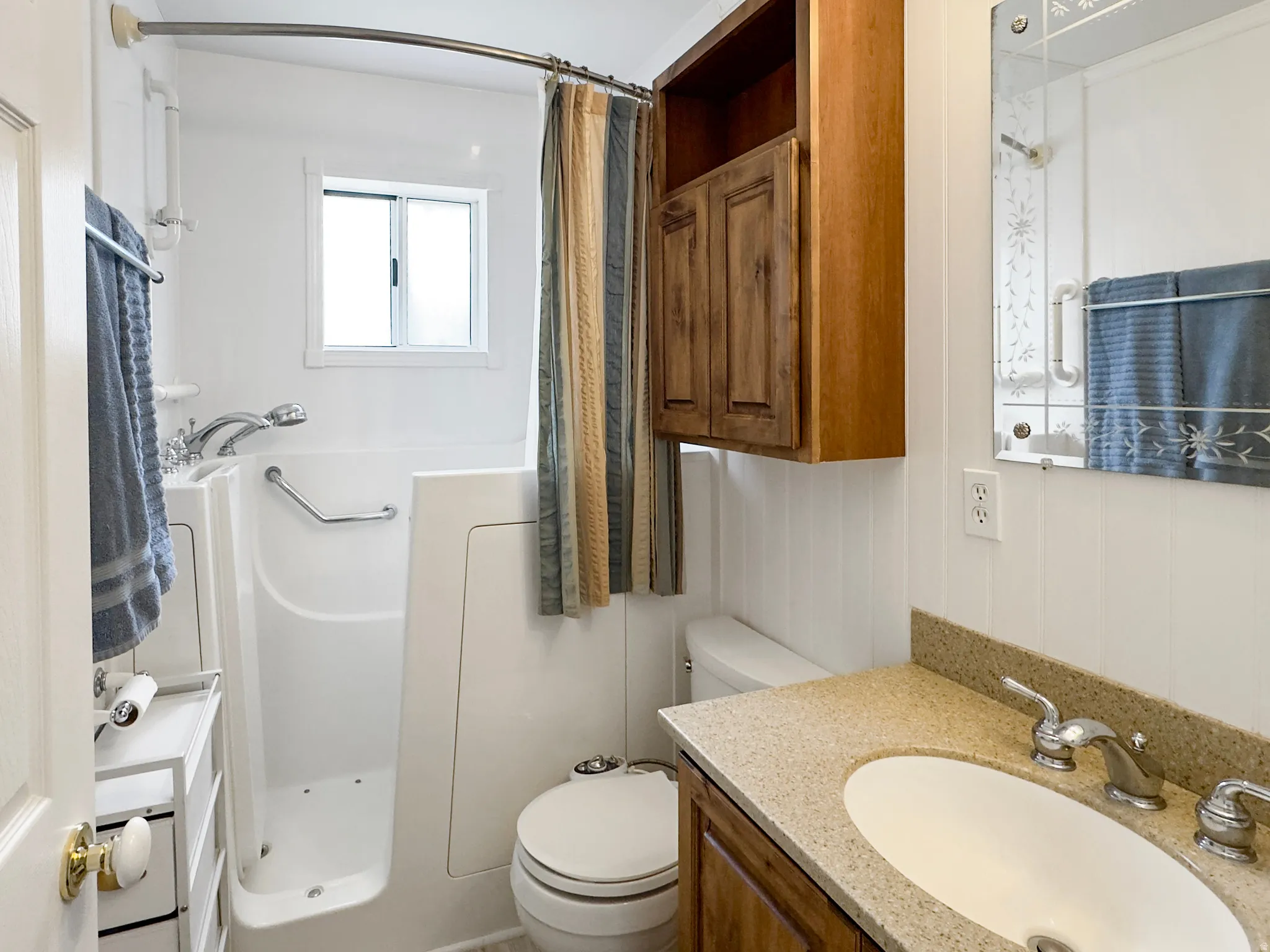 White bathroom featuring a walk-in tub with grab bars, a window, and a wood-finish vanity with a light-toned countertop and an oval sink