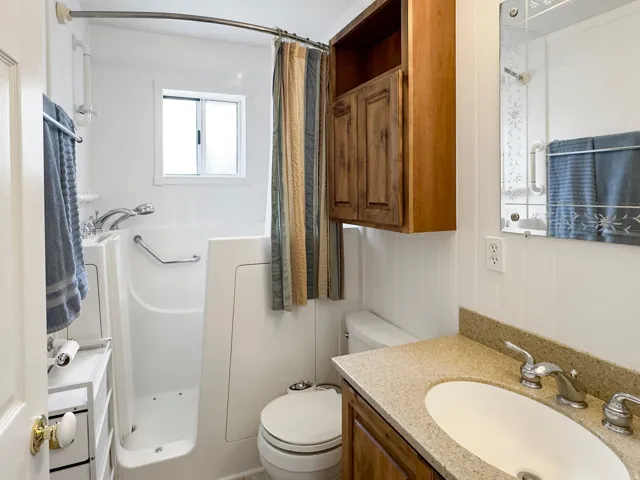 White bathroom featuring a walk-in tub with grab bars, a window, and a wood-finish vanity with a light-toned countertop and an oval sink