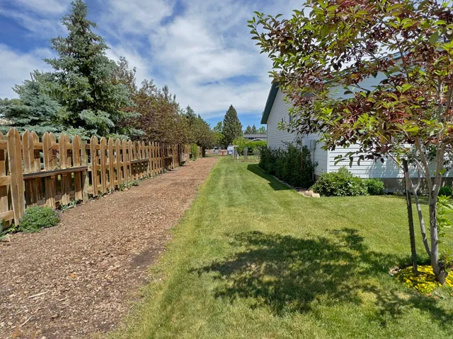 Expansive side yard featuring a long stretch of green lawn, a wooden picket fence, mature trees, and a gravel pathway