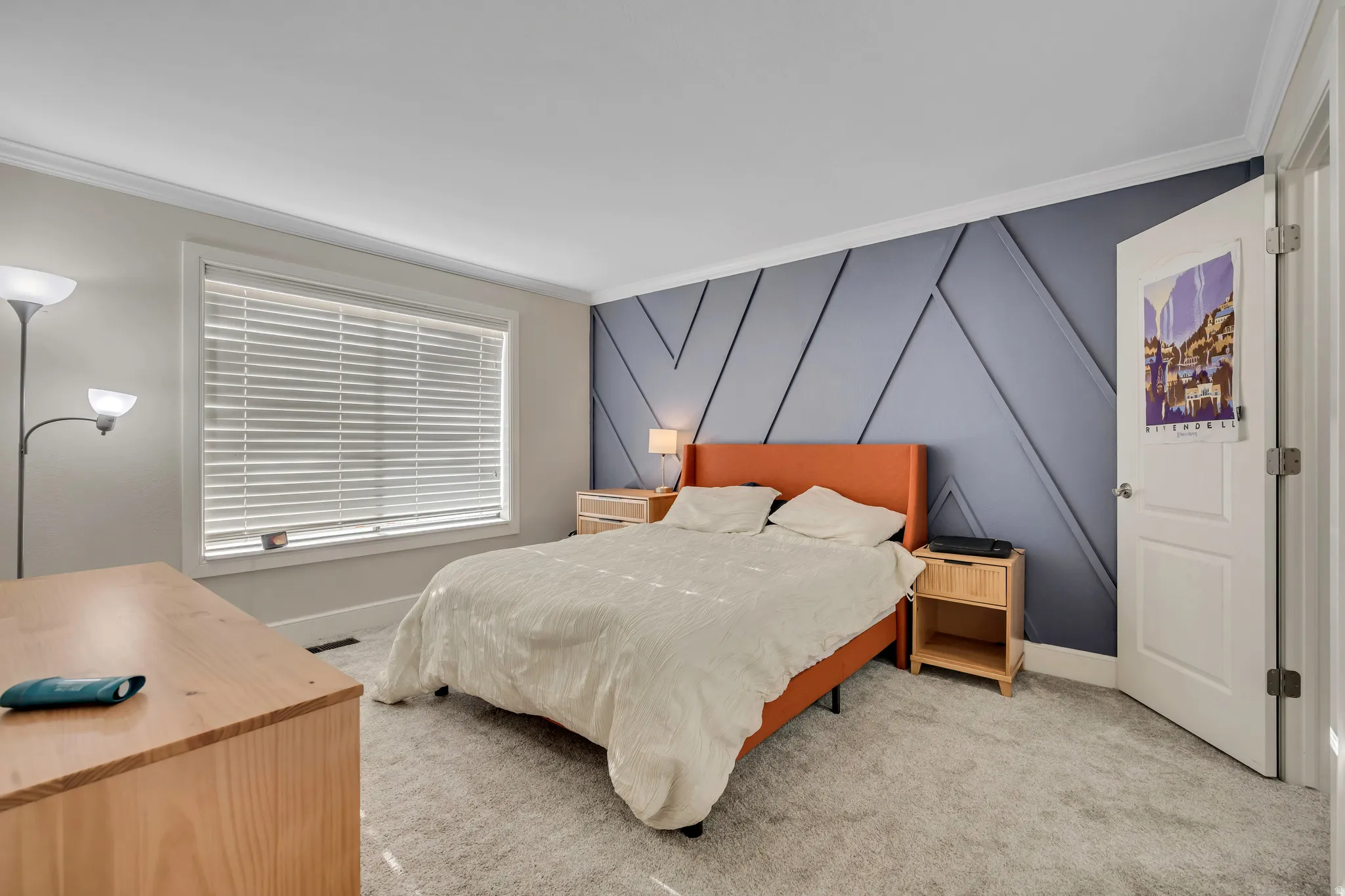 Bedroom featuring a decorative accent wall with geometric paneling, natural light from a window with blinds, light-toned carpeting, and white trim