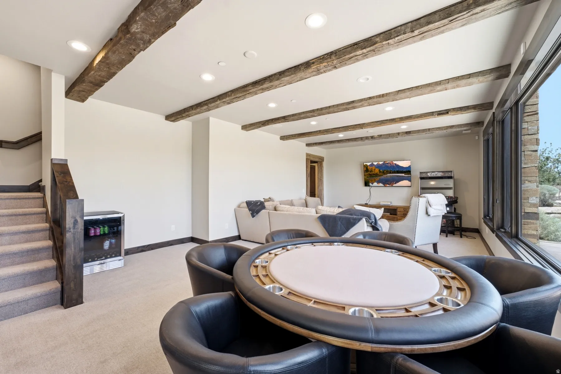 Carpeted lower-level room featuring exposed wood ceiling beams, recessed lighting, and a beverage cooler