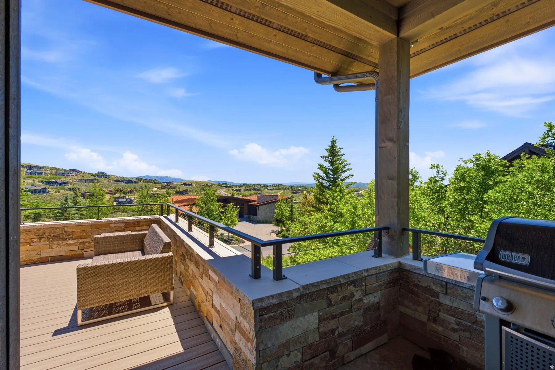 Covered outdoor deck featuring composite decking, stone veneer walls, black metal railings, and an expansive distant landscape